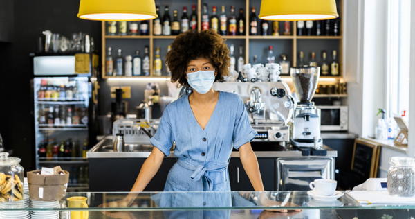 Young female owner wearing mask standing at counter in coffee shop